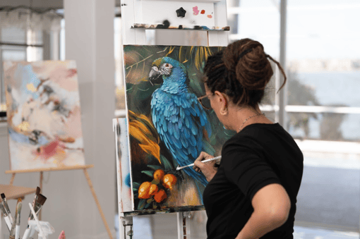 woman painting destils on canvas of blue bird and orange flowers